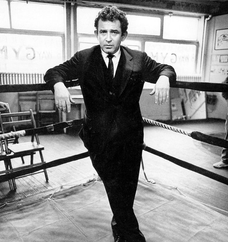 Norman Mailer in a suit and tie, leaning back against the ropes of a boxing ring in a gym, about 1955